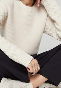 Lichtbeige gebreide trui met een gestructureerd patroon op de mouwen, geribbelde boorden, ronde halslijn, gecombineerd met zwarte broek en witte sneakers.