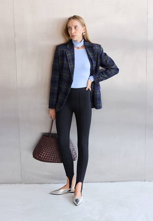 Žena u plavoj majici i kariranom blejzeru stoji uz zid držeći smeđu pletenu torbu, nosi crne hlače i srebrne cipele s špicom.