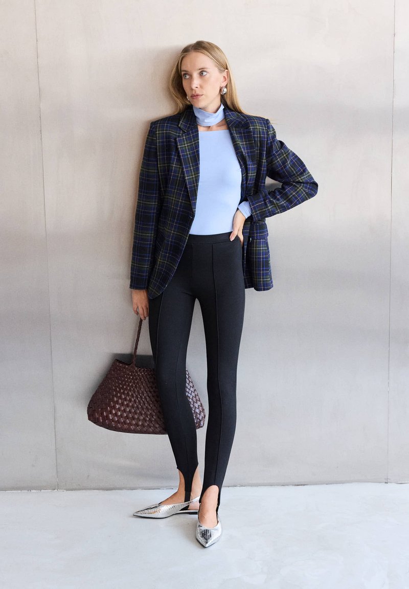 Γυναίκα με μπλε τοπ και καρό σακάκι στέκεται στον τοίχο κρατώντας καφέ πλεκτή τσάντα, φορά μαύρο παντελόνι και ασημένια μυτερά μπαλαρίνια.