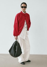Femme portant des lunettes de soleil noires, une veste rouge, une chemise blanche boutonnée et un pantalon blanc large, tenant un grand sac noir, debout sur un sol beige.
