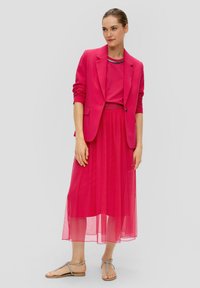 Felroze blazer over een bijpassende top, gecombineerd met een sheer roze rok en zilveren sandalen. De outfit heeft een gedetailleerde halslijn met kralen.