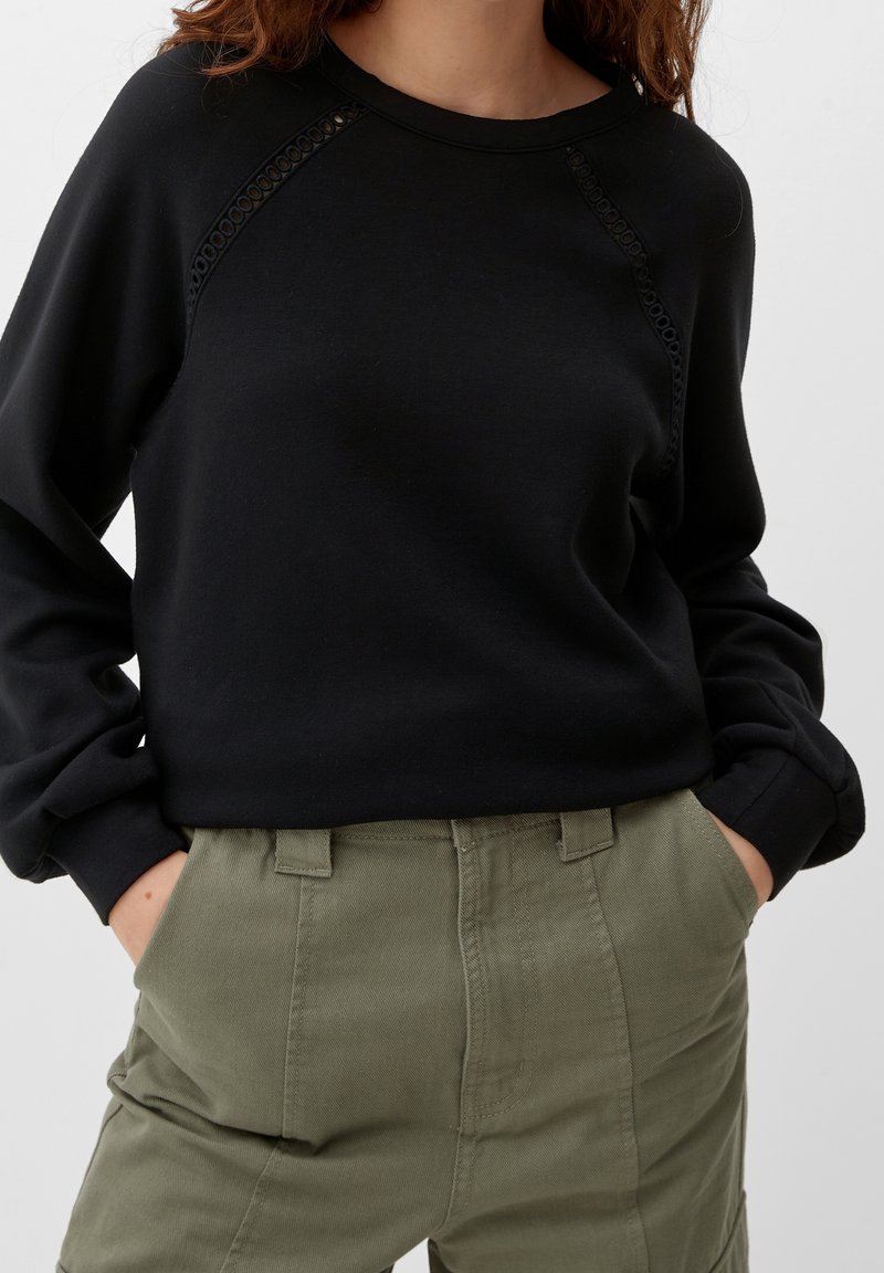 Femme portant un haut noir à manches longues avec un détail en dentelle sur les épaules et un pantalon taille haute vert olive, les mains dans les poches.