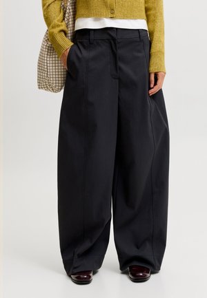 Personne portant un pantalon noir à jambes larges, un cardigan moutarde sur une chemise blanche, tenant un sac à carreaux et portant des chaussures marron foncé.