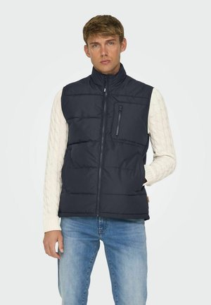Gilet imbottito blu navy con colletto alto, design a righe orizzontali, tasca con zip frontale e tasche laterali, indossato sopra un maglione bianco a maglia cable.
