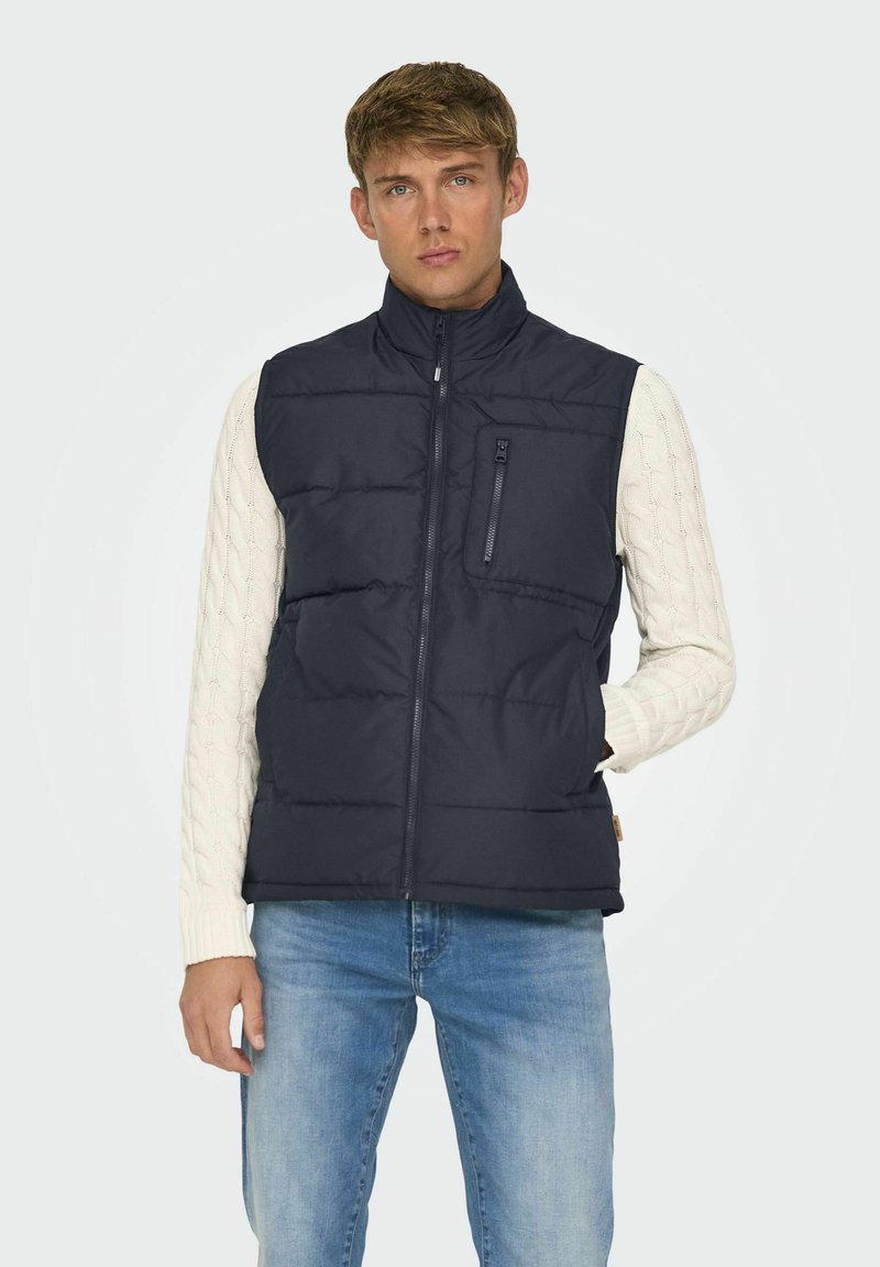 Veste sans manches rembourrée bleu marine avec col montant, design matelassé horizontal, poche zippée à l'avant et poches latérales, portée par-dessus un pull en maille torsadée blanche.
