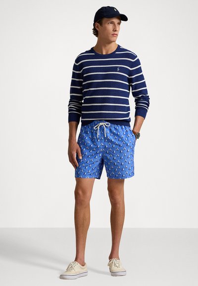 Marine stripete genser, blå badshorts med seilbåt mønster, og beige sneakers. Tilbehøret inkluderer en marineblå caps og et armbåndsur.