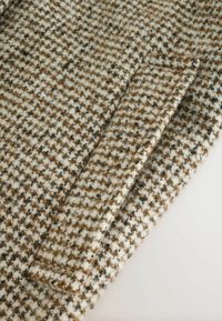 Tissu en laine au motif pied de poule avec des couleurs marron, beige et noir. Il présente une surface texturée et un détail de bord replié.