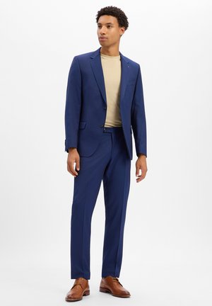 Ein Mann trägt einen blauen Anzug mit einem beigen Hemd und braunen eleganten Schuhen und steht vor einem schlichten weißen Hintergrund.