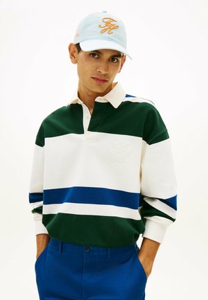 Joven con una camiseta de rugby a rayas en verde, blanco y azul, pantalones azules y una gorra azul clara con letras bordadas en amarillo.