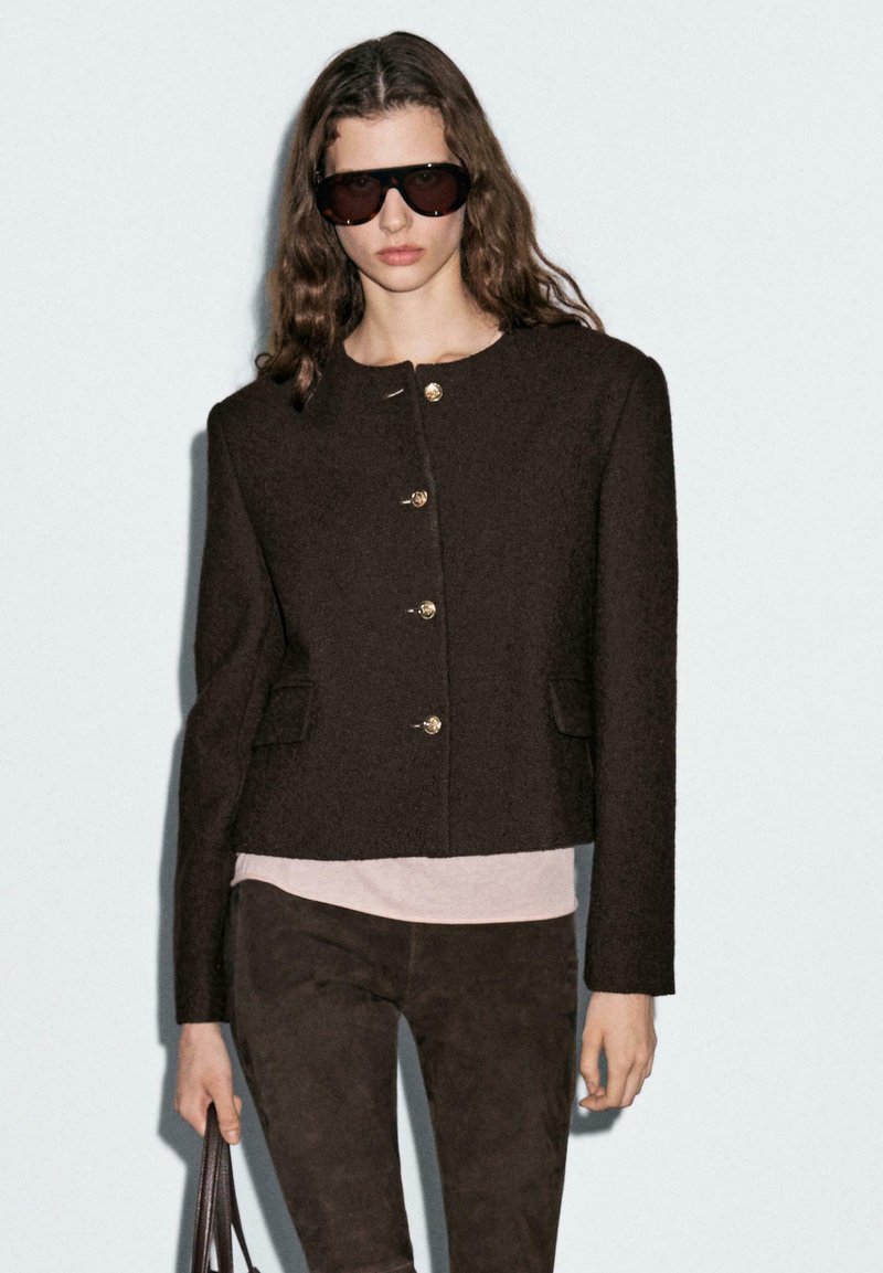Brown cropped jacket with gold buttons, textured fabric, and side pockets, paired with a light pink top and dark brown pants.