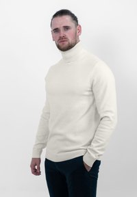 Crème coltrui gemaakt van zacht breigaren. Kenmerken zijn fitted lange mouwen, ribbelboorden en -zoom, een gladde textuur en een naadloos ontwerp.