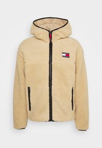 Giacca a vento beige in sherpa con cappuccio, zip nera, tasche laterali e patch con logo Tommy Jeans sul petto.