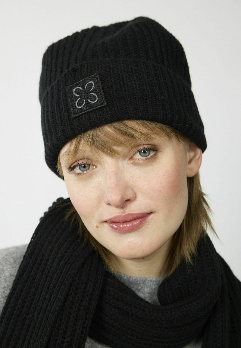 Zwarte geribde gebreide beanie met een vierkante patch met een grijs ontwerp. Gepaard met een bijpassende zwarte sjaal, beide gemaakt van zacht materiaal.