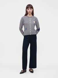 Femme portant un cardigan rayé boutonné avec des patches, un pantalon large foncé et des ballerines pointues noires, debout les mains derrière le dos.