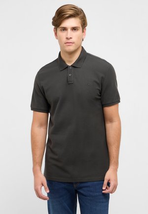 Młody mężczyzna o jasnobrązowych włosach, ubrany w ciemnoszarą koszulkę polo i niebieskie dżinsy, stojący na tle jednolitego jasnego tła.