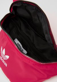 Röd nylonpåse med dragkedja, svart insida, med en vit Adidas-logotyp på utsidan och en etikett inuti. Kompakt design för enkel förvaring.