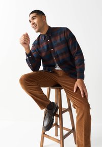 Koszula zapinana na guziki w brązowo-niebieską kratę, zestawiona z brązowymi spodniami sztruksowymi i czarnymi butami, siedząca na drewnianym stołku na białym tle.