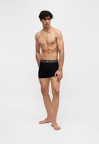 Jeune homme debout pieds nus portant un boxer noir avec une ceinture dorée à motif sur un fond uni.