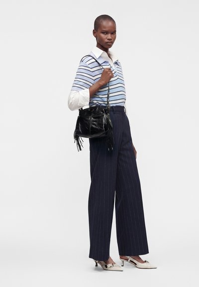 Femme portant un pull bleu rayé sur une chemise blanche, un pantalon large à fines rayures sombre, des talons blancs et tenant un sac à main en cuir noir à franges.