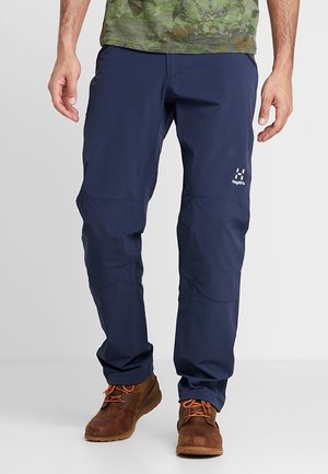 Mężczyzna w granatowych spodniach outdoorowych i brązowych sznurowanych butach, stojący na jednolitym tle.