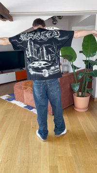 Must värviline T-särk, millel on linnavaade, auto ja ämblikuvõrgu kujundus. Seljas sinised teksad, seismine toas, kus on taimi ja mööblit.