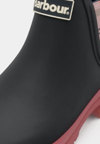 Μαύρη λαστιχένια βροχή μπότες με κόκκινη σόλα, που διαθέτει λευκή ετικέτα με λογότυπο και καρό λεπτομέρεια στο επάνω μέρος, λεία υφή και στρογγυλεμένο σχήμα.