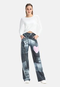 Sieviete, kura ģērbusies baltā ar garām piedurknēm topā un plašu kāju biksēs ar džinsa patchwork rakstu un rozā sirds dizainu, rokās ielikusi kabatās.