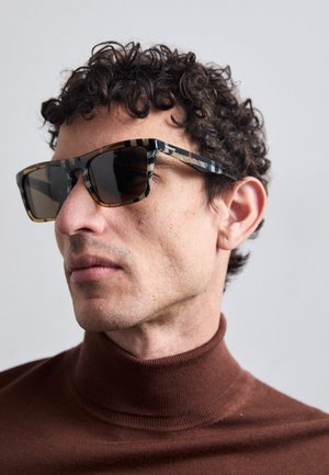 Gafas de sol con forma rectangular y montura negra y carey con un patrón, que cuentan con lentes oscuros. Llevadas por una persona con cabello rizado.