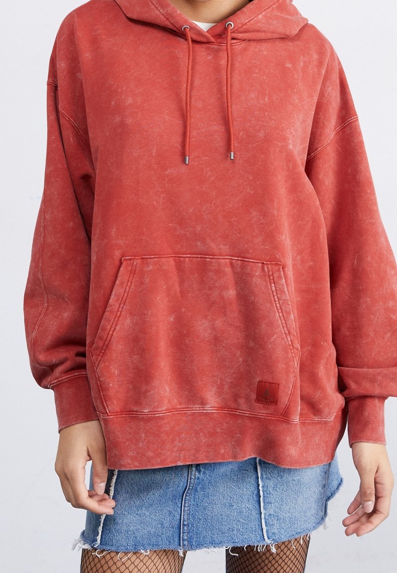 Sudadera roja oversized con acabado texturizado, cordones, bolsillo delantero y parche con logotipo. Llevada sobre una falda vaquera con dobladillo deshilachado.