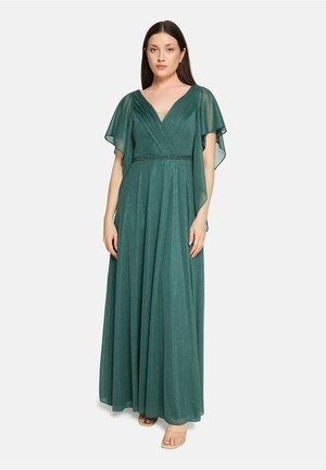 Kvinde iført gulvlang smaragdgrøn glitrende kjole med V-hals og flagrende ærmer, stående mod en ensartet lys baggrund.