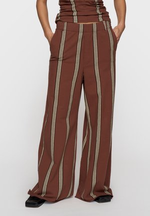 Brązowe spodnie o szerokich nogawkach z beżowymi pionowymi paskami, noszone z dopasowaną bluzką i czarnymi butami na kwadratowym nosku, ręce w kieszeniach.