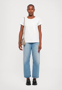 White cotton t-shirt with large “AX” graphic, paired with light blue straight-leg jeans and black loafers. Beige crossbody bag.