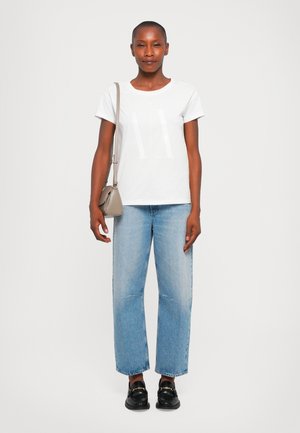 T-shirt en coton blanc avec un grand graphique "AX", associé à un jean droit bleu clair et des mocassins noirs. Sac en bandoulière beige.