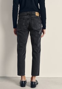 Oseba, oblečena v črne ozke kavbojke, črno majico z dolgimi rokavi in črne loaferje, stoji obrnjen proti svetli enobarvni podlagi.