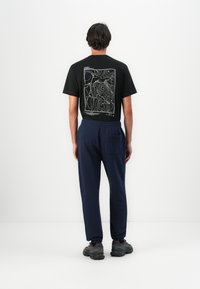 Mann iført svart t-skjorte med hvit konturkartdesign på ryggen, marineblå joggebukser og svarte joggesko som står mot hvit bakgrunn.