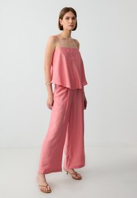 Roze, mouwloze top met een uitlopende vorm, gecombineerd met ruime roze broeken. Zacht stof, minimale naden en een subtiele glans. Eenvoudige sandalen met bandjes.
