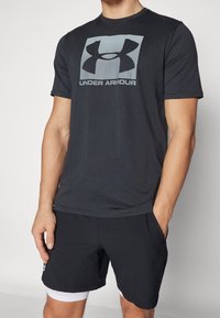 Camisa negra de manga corta con el logo gris de Under Armour, combinada con pantalones cortos negros. El material parece suave y ligero. Diseño sencillo.
