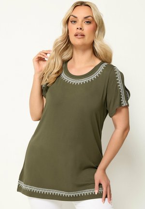 Lyshåret kvinde iført olivengrøn tunika med hvid geometrisk broderi på halsudskæring, ærmer og kant, poserer mod hvid baggrund.