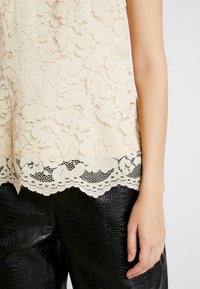 Beige blommig spets topp med vågig nederkant i kombination med svarta texturerade faux l äderbyxor, visar höger arm och hand avslappnad vid sidan.