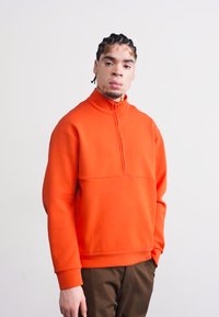 Pullover laranja com fecho de quatro partes, feito de tecido suave, apresentando uma costura frontal e um corte descontraído. Usado com calças castanhas.