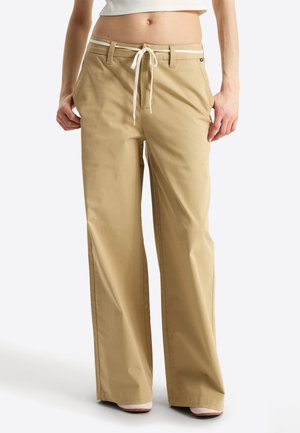 Beige weite Hosen mit Knopf, Gürtelschlaufen und weißem Kordelzug, getragen von einer Person mit kurzem weißen Top vor weißem Hintergrund.
