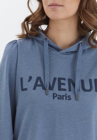 Ljusblå hoodie med tryck på framsidan, dragsko med metallspetsar och mjukt, texturerat material.