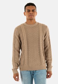 Maglione beige lavorato a maglia con motivo a trecce, scollo rotondo, polsini e orlo a costine. Indossato sopra jeans azzurri chiari.