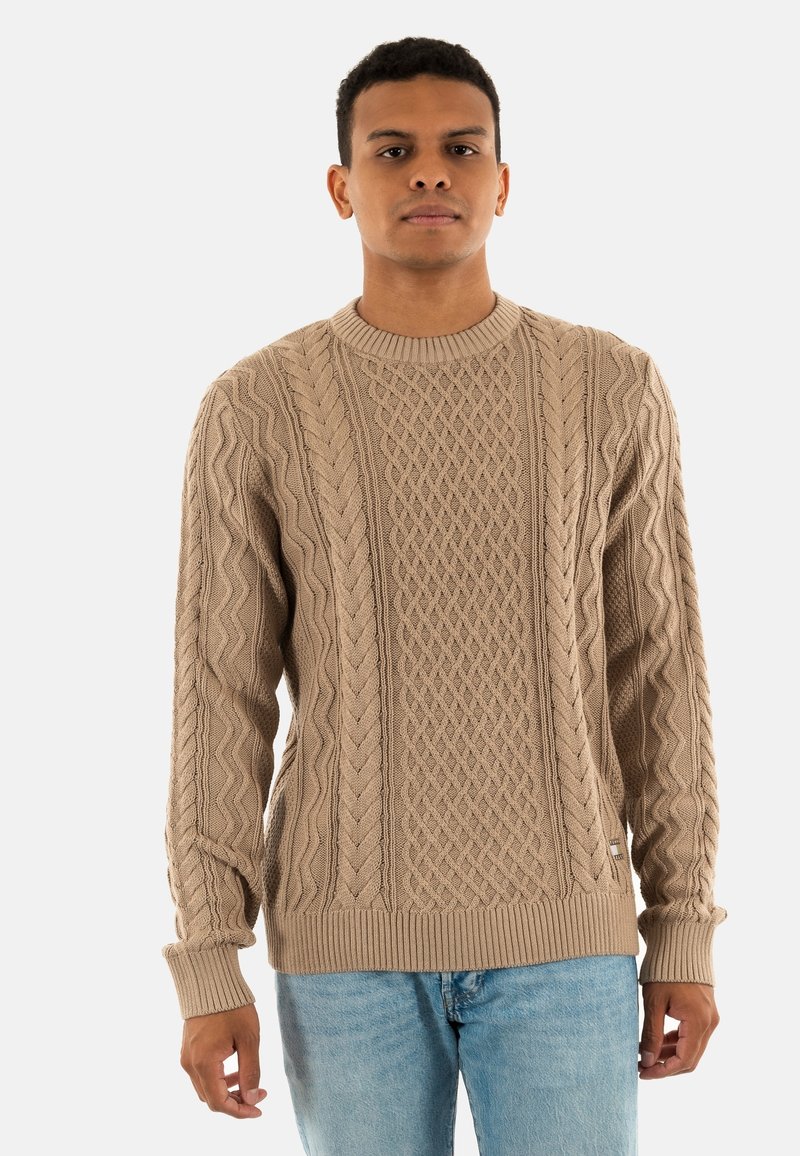 Maglione beige lavorato a maglia con motivo a trecce, scollo rotondo, polsini e orlo a costine. Indossato sopra jeans azzurri chiari.
