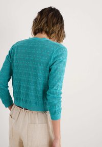 Tiffany sininen neulecardigan, jossa on teksturoitu kuosi, pitkät hihat ja lyhyt malli, yhdistettynä vaaleisiin pellavahousuihin.