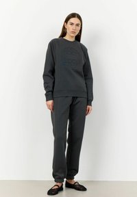 Sweatshirt gris foncé avec logo en relief, associé à un pantalon de jogging assorti avec des poignets élastiques. Le modèle se tient sur un fond neutre.