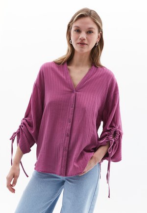 Blusa rosa, strutturata, con scollatura a V, chiusura a bottoni e maniche leggermente arricciate con dettagli a nodo. Abbinata a jeans azzurri chiari.