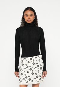 Žena s kovrčavom kosom nosi crnu bluzu s dugim rukavima i visokim ovratnikom te bijelu suknju s crnim cvjetnim uzorkom, stoji na bijeloj pozadini.