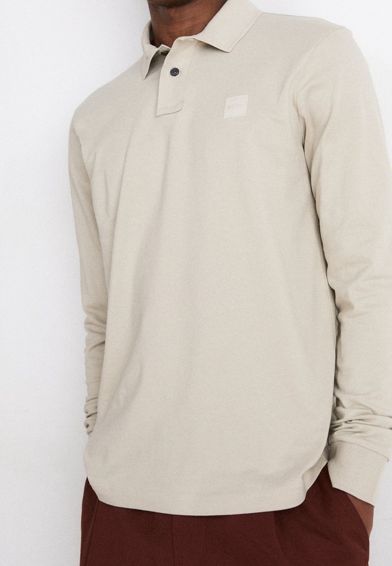Beige longsleeve poloshirt met een kraag en twee zwarte knopen. Voorzien van een klein logo-op-label op de borst. Zachte, gladde textuur.