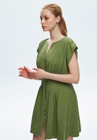 Grünes Leinenkleid mit kurzen Ärmeln, durchgehender Knopfleiste und vertikalen Falten, mit einem leichten Schlitz am Saum für Details und Textur.